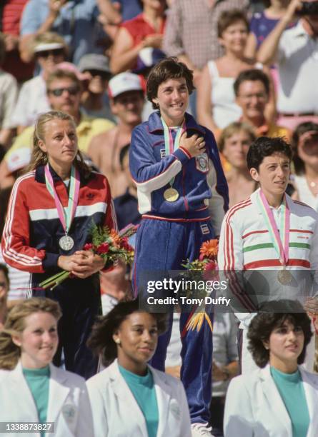 Joan Benoit of the United States stands on the podium during the playing of the national anthem beside silver medallist Grete Waitz of Norway and...