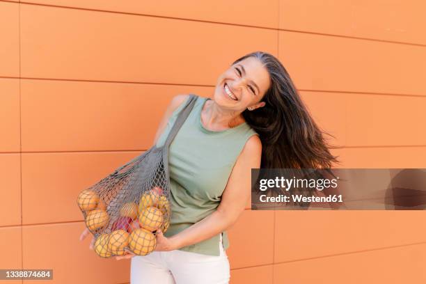 happy woman with mesh bag of fruits standing by orange wall - tossing hair facing camera woman outdoors stock-fotos und bilder