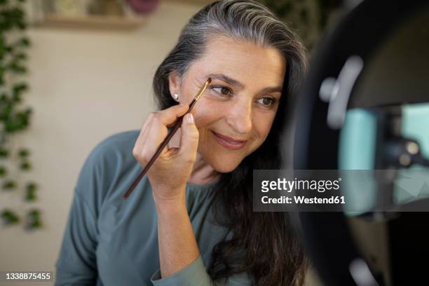 smiling female vlogger brushing eyebrows during tutorial at home - eyebrow stock-fotos und bilder