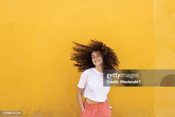 carefree woman tossing hair in front of yellow wall - hair toss stock pictures, royalty-free photos & images