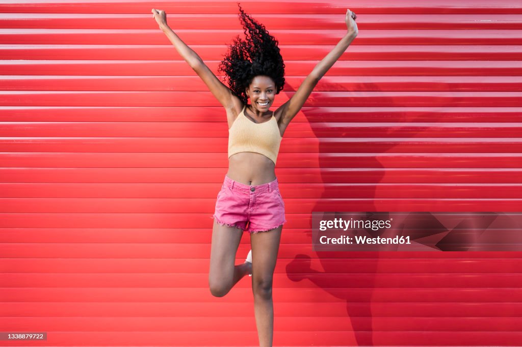 Happy young woman with arms raised jumping in front of red shutter