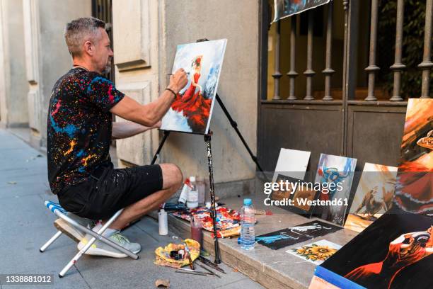 male painter painting on canvas while sitting on street - street artist - fotografias e filmes do acervo