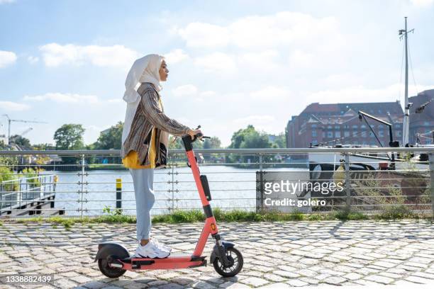 young woman with hijab on e-scooter at riverside in modern berlin - hijab fashion stockfoto's en -beelden