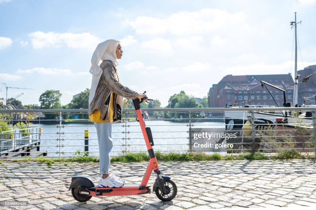 Young woman with hijab on e-scooter at riverside in modern berlin