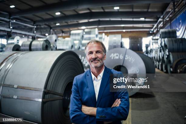 smiling businessman with arms crossed standing at warehouse - metallindustrie stock-fotos und bilder