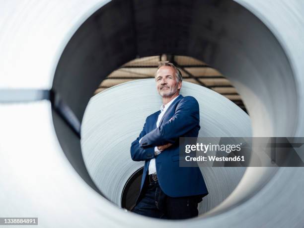 confident businessman with arms crossed standing by metal sheet roll - männerrunde stock-fotos und bilder