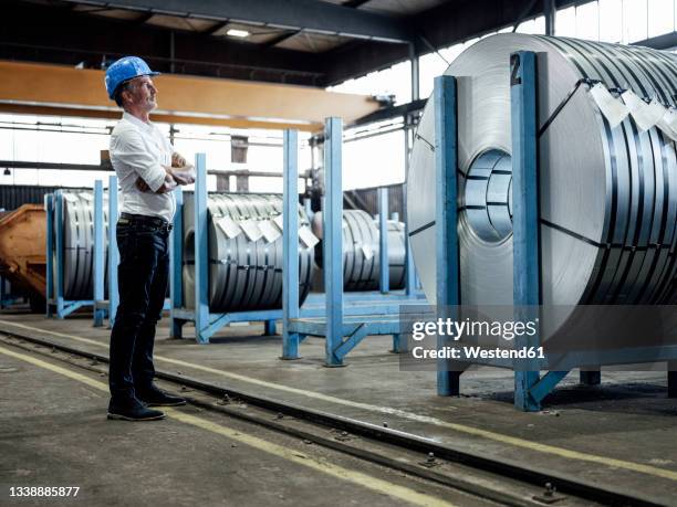 male engineer with arms crossed checking metal sheet roll in steel mill - industria pesada fotografías e imágenes de stock