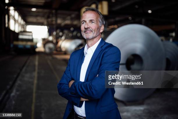 thoughtful businessman with arms crossed standing at metal industry - three quarter length stock pictures, royalty-free photos & images