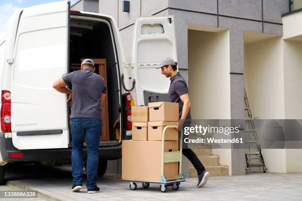 man opening door while colleague pushing cart with boxes near van - moving truck stock pictures, royalty-free photos & images