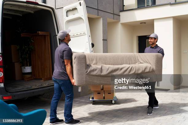 male delivery coworkers unloading sofa from moving van near house - camión de las mudanzas fotografías e imágenes de stock