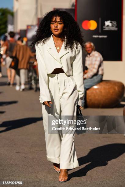 Passerby wears silver earrings, a gold chain pendant necklace, a white latte oversized cropped blazer jacket, matching white latte high waist flared...