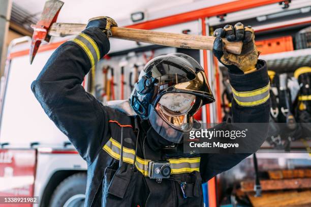 side view of unrecognizable firefighter in uniform and hardhat carrying big used pickax with wooden handle while standing near fire truck with professional tools and safety vests - fireman axe stock pictures, royalty-free photos & images