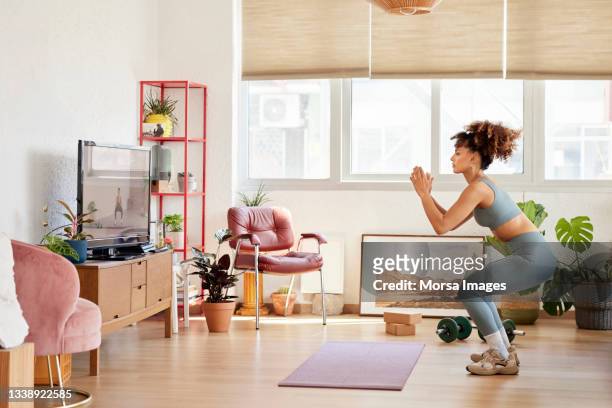 young woman practicing squats in living room - crouching ストックフォトと画像