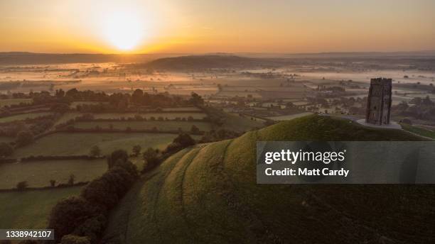 The sun rises over mist that has formed on the Somerset Levels beyond St. Michael's Tower at the top of Glastonbury Tor in the town of Glastonbury,...