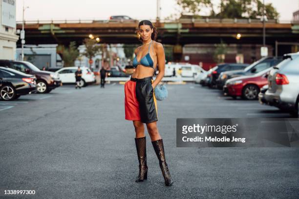 Model Nakisa Kachingwe wears a blue denim bikini top, red and black leather shorts, black see-through heeled boots, and carries a small baby blue...