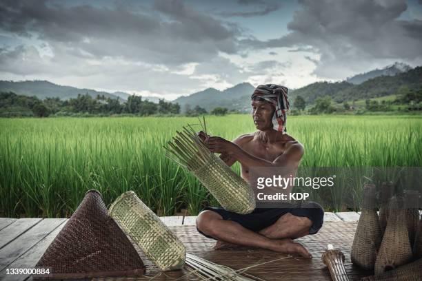thai lifestyles and bamboo handicraft, asian mature man is making fishing traps by bamboo weaving in his cottage with agriculture rice fields  background. thai basketry hand craft - daily life traditional fishermen stock pictures, royalty-free photos & images