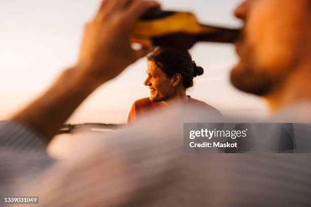 man drinking beer while smiling male friend looking away at lakeshore in evening - men friends beer outside stock-fotos und bilder