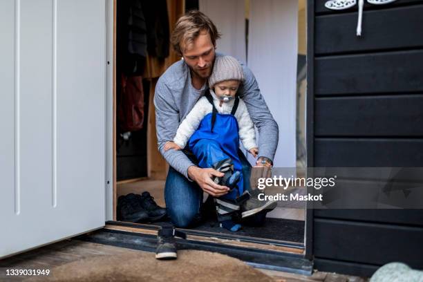 mid adult man helping son put on shoe at doorway - getting dressed stockfoto's en -beelden