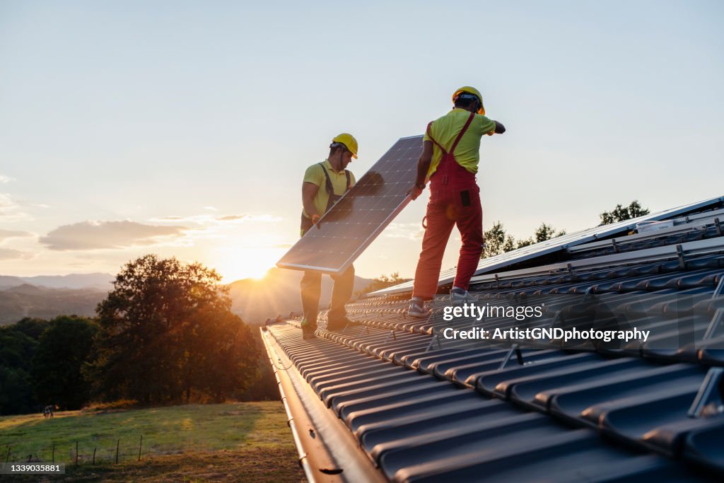 Arbeiter, die Sonnenkollektoren auf einem Holzhaus in der Natur bei Sonnenuntergang installieren.