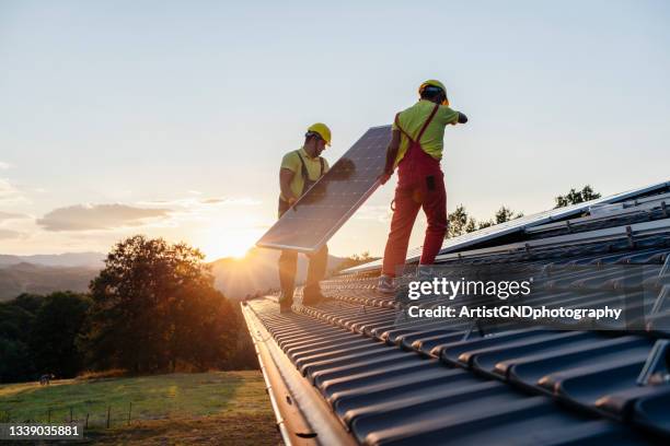 arbeiter, die sonnenkollektoren auf einem holzhaus in der natur bei sonnenuntergang installieren. - dach stock-fotos und bilder