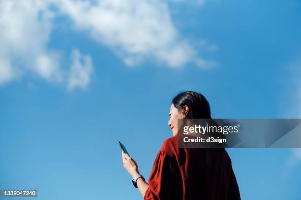 low angle portrait of young asian woman using smartphone against beautiful blue sky with cloudscapes, enjoying sunlight outdoors. lifestyle and technology - erwartung stock-fotos und bilder