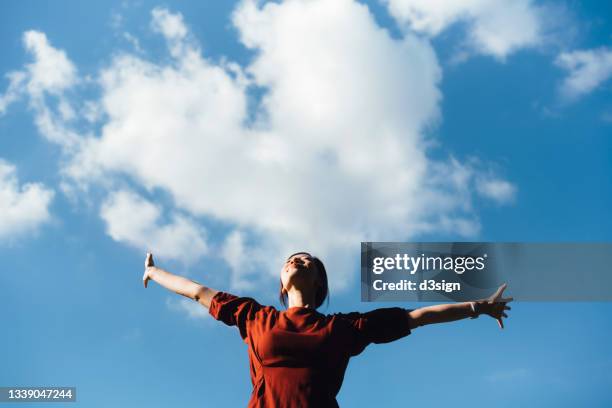 low angle portrait of young asian woman with her eyes closed stretching arms, setting herself free and feeling relieved. enjoying fresh air and sunlight with head up against beautiful blue sky with cloudscapes. freedom in nature. connection with nature - arms outstretched foto e immagini stock