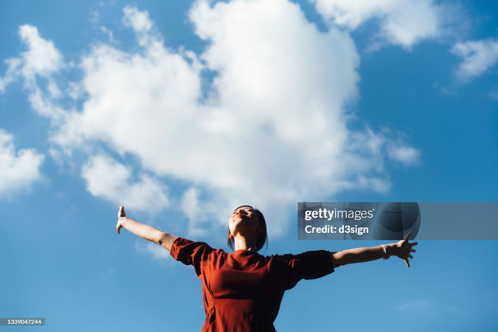 Low angle portrait of young Asian woman with her eyes closed stretching arms, setting herself free and feeling relieved. Enjoying fresh air and sunlight with head up against beautiful blue sky with cloudscapes. Freedom in nature. Connection with nature