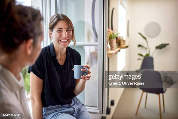 two businesswomen having a coffee break at the window in office - coffee break fotografías e imágenes de stock