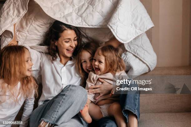 happy mother with three girls holding blanket on sofa at home - home family stockfoto's en -beelden