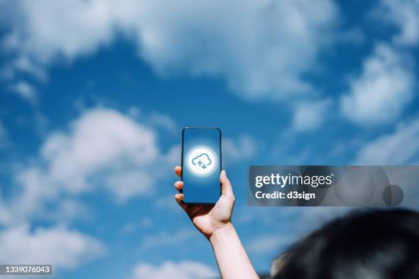 close up of woman's hand raised holding smartphone against beautiful blue sky with cloudscapes, with cloud computing icon on the device screen. the delivery of data storage, servers, databases, networking and softwares through the internet concept - mobile phone evolution stock-fotos und bilder