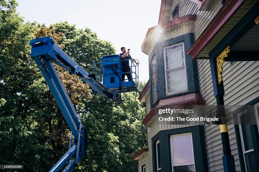 Mature man standing on cherry picker power washing home exterior
