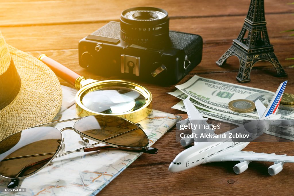 Items For Summer Holidays: A Camera, Sunglasses, Cash, A Straw Hat, A Map And A Travel Plan. Tourist layout-set and accessories of the Traveler, On a wooden background. Open borders concept for free happy travel. Copy of the text space.