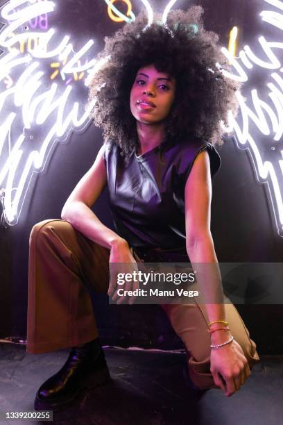 afro american woman crouching looking at camera with a white glowing neon wings sign behind on wall - dark angel stock pictures, royalty-free photos & images