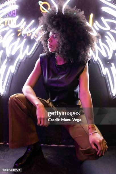 afro american woman crouching looking away with a white glowing neon wings sign behind on wall - disco costume stockfoto's en -beelden