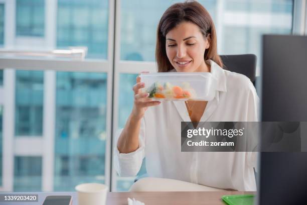 femme souriante à son bureau sentant les légumes dans une boîte en plastique pour le déjeuner - odorat photos et images de collection