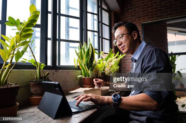 senior asian business executive having take away food during lunch break at eco-friendly cafeteria with beautiful potted plants decoration. the mature businessman surfing net and checking mails using laptop and sitting next to balcony window. - smart casual lunch stock pictures, royalty-free photos & images