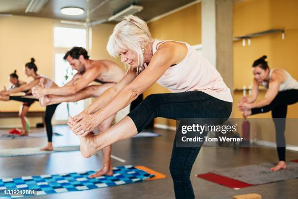 mature woman holding foot stretched in front of her during yoga class - hot yoga stock pictures, royalty-free photos & images