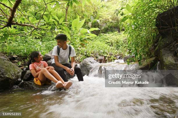 brother and sister sitting on a rock and playing in a stream of water - indian family vacation stock pictures, royalty-free photos & images