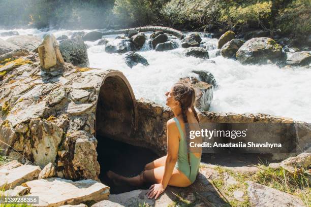 young woman traveler relaxing at the natural hot spring by the mountain river in kyrgyzstan during sunny day - kyrgyzstan stock pictures, royalty-free photos & images