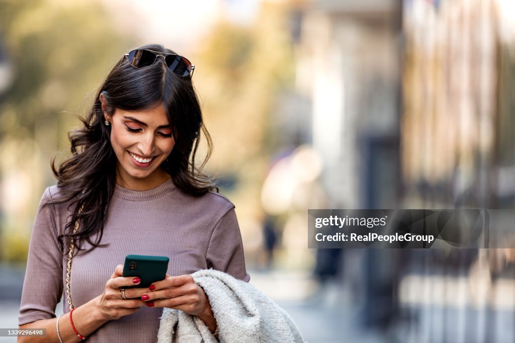 Beautiful mid adult woman walking and texting message on mobile phone outside business center.