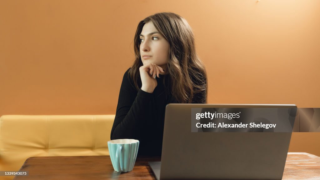 Portrait of young girl on yellow background with copy space. Concept with hot beverage and gray laptop. She is drinking a coffee and working on computer
