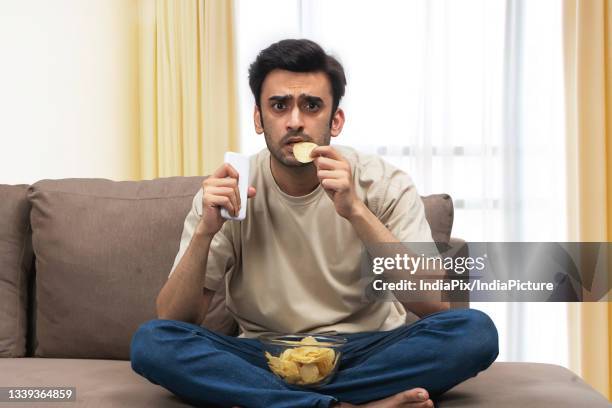 a young man with remote sitting tensed on sofa eating chips while watching sports. - sports india stockfoto's en -beelden