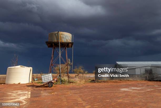 stormy dark sky's in the background of a old farm  shearing shed and water tank with red dirt and sunlight in the foreground, outback australia - australia storm stock pictures, royalty-free photos & images