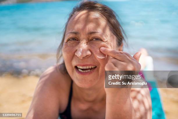 ältere frau, die sonnencreme auf ihr gesicht auftrauft und lächelt, während sie sich am strand sonnt - sunburn tourist stock-fotos und bilder