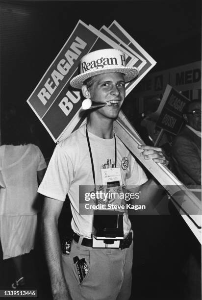 Portrait of an unidentified young man, a noisemaker in his mouth, a straw hat on his head, and collection of sign endorsing 'Reagan; Bush,' as he...