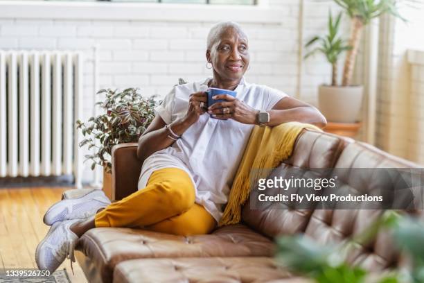 beautiful senior woman smiling while sitting on sofa and drinking tea at home - middle aged woman at home stock pictures, royalty-free photos & images