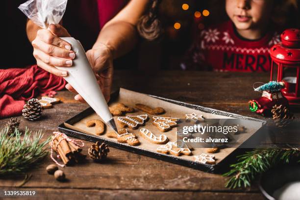 woman icing gingerbread christmas cookie by son in kitchen - fazer doces imagens e fotografias de stock