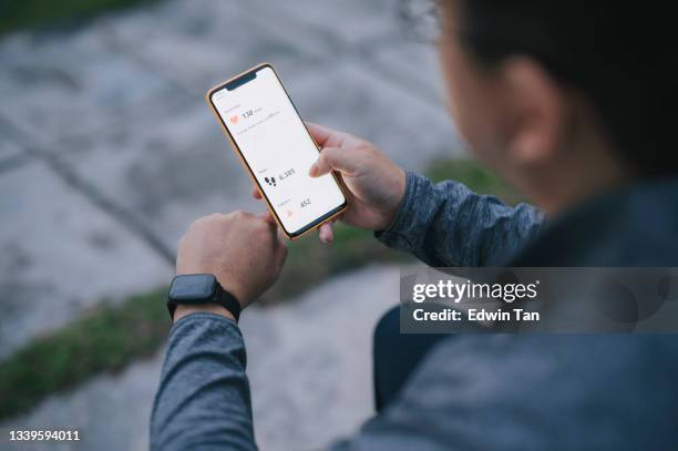 over shoulder view asian chinese man using fitness tracker mobile app connecting to fitness tracker at public park after exercise in the morning - fit man stockfoto's en -beelden