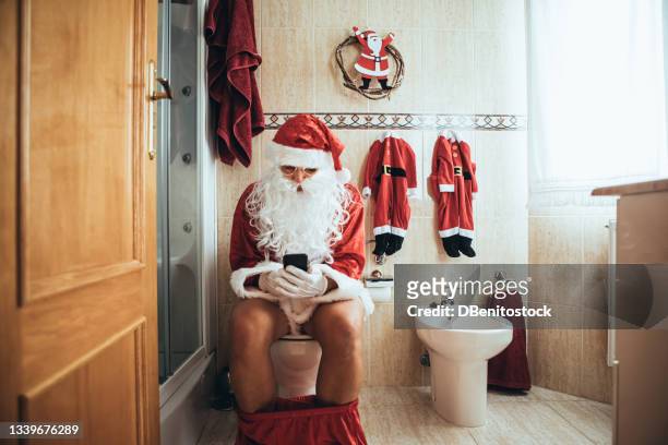 santa claus at christmas, in his bathroom, sitting on the toilet, looking at his mobile - schocktaktik stock-fotos und bilder