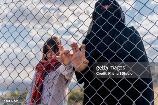 refugee woman and daughter standing behind a fence - refugee camp stock pictures, royalty-free photos & images